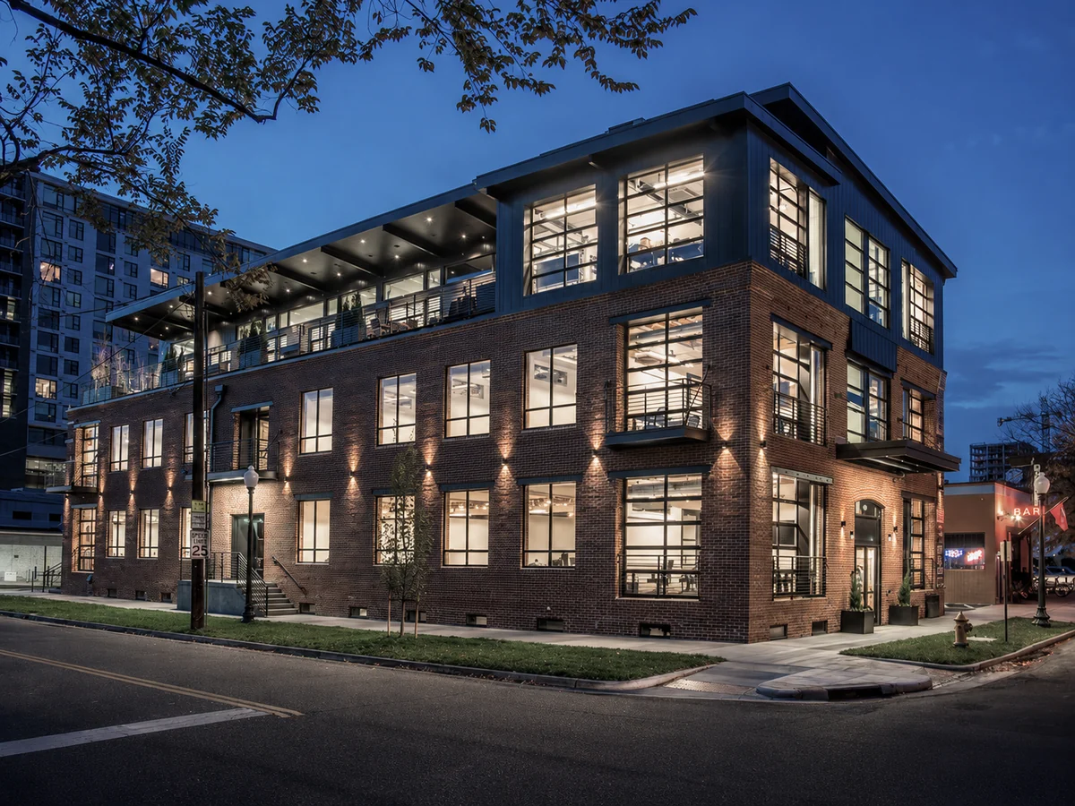 The Company Sellers office at 1001 Bannock Street, Denver &mdash; a three-storey red brick corner building with black steel windows, lit interiors and a covered rooftop terrace, photographed at dusk